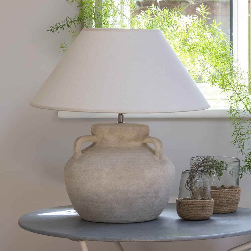 Table lamp with beige base and white shade on a table with plants in the background