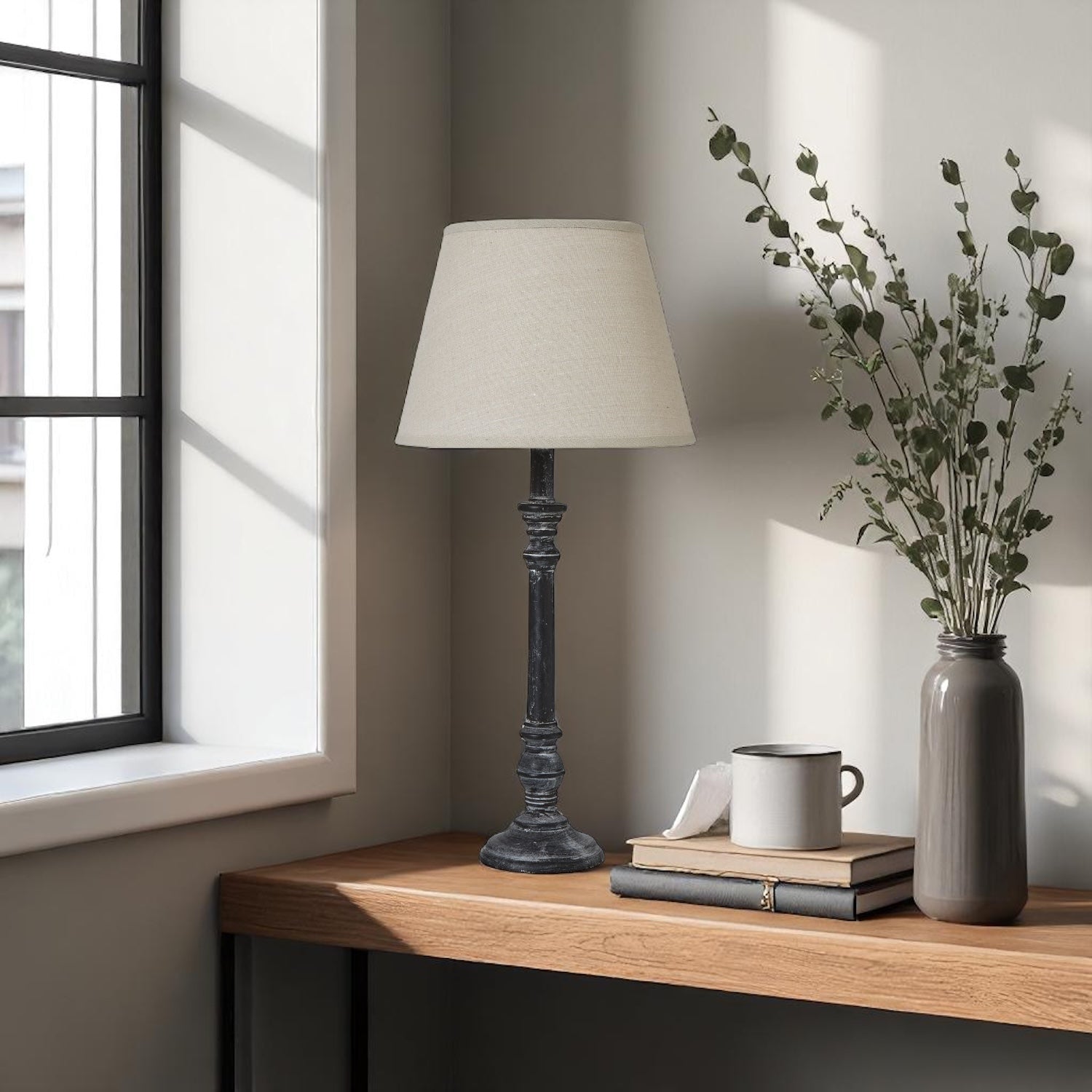 Table lamp, mug, books, and vase on a wooden surface near a window.