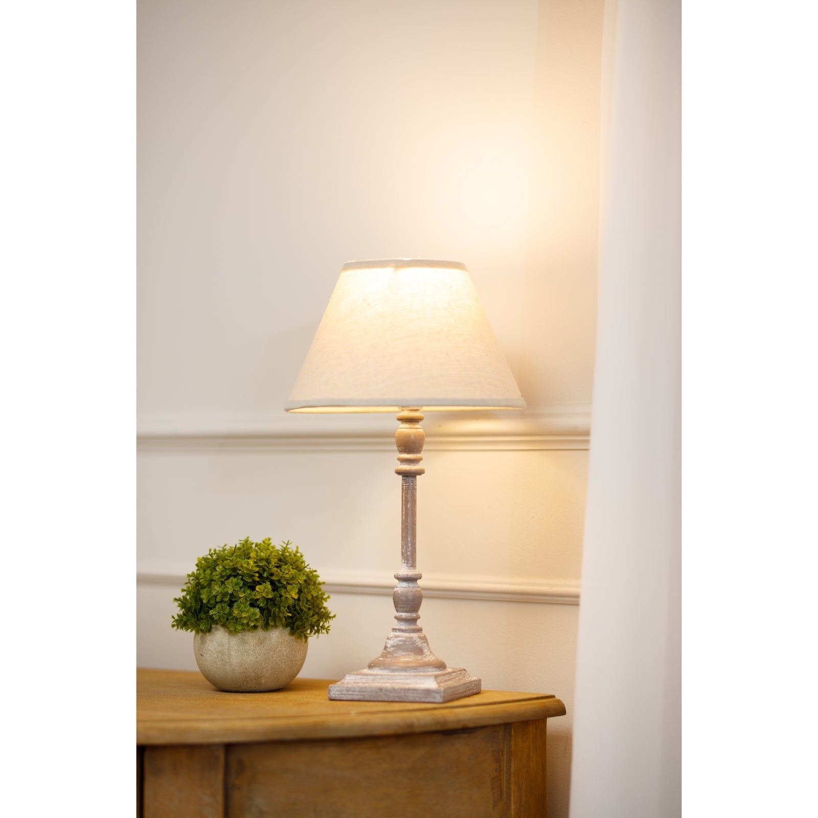 Almira Table lamp with a white shade on a wooden table next to a small potted plant, against a light-coloured wall.