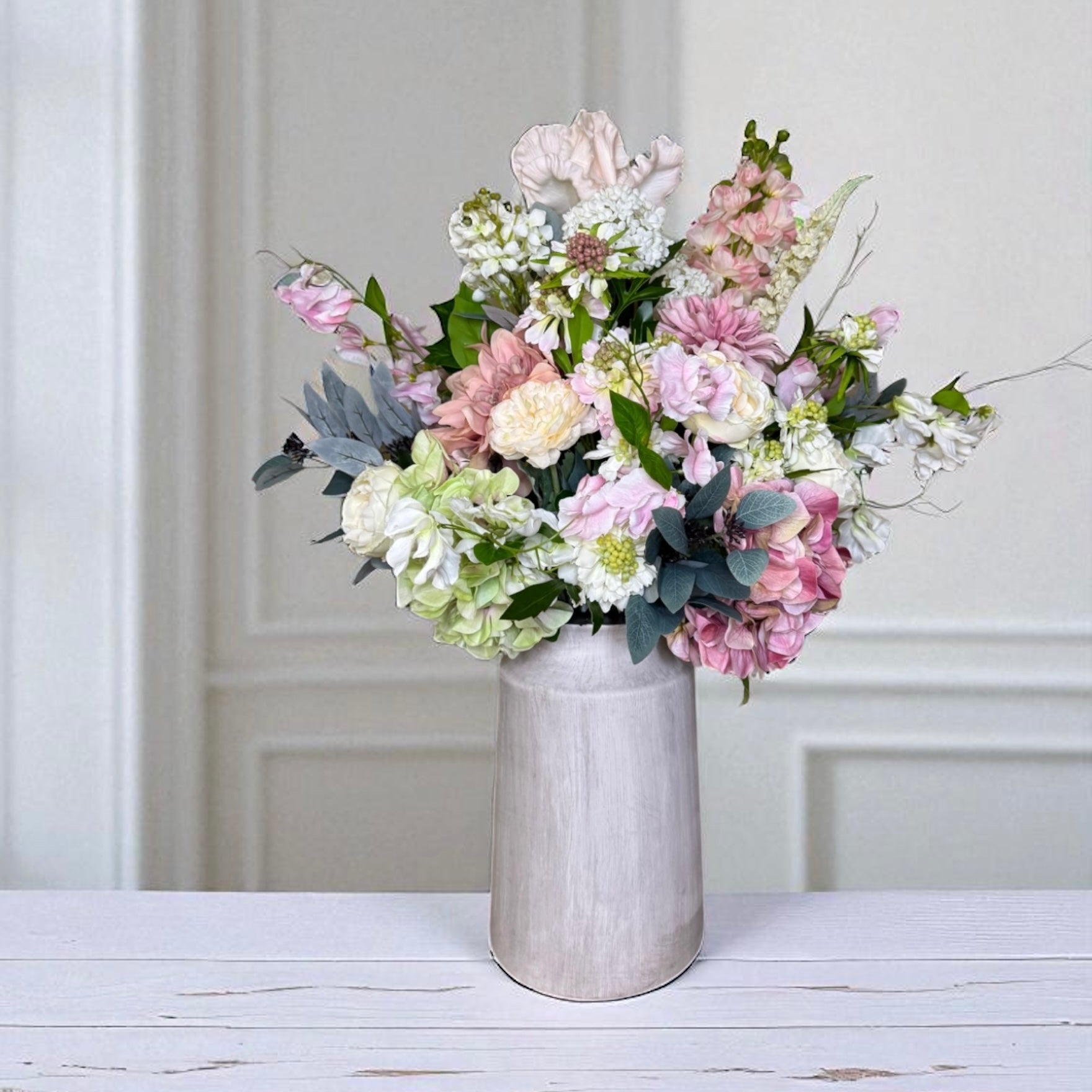 Large bouquet of pink, white and green flowers in a vase on a wooden table with a white panelled wall background