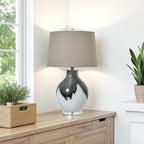 Grey and white glass table lamp with a beige shade on a wooden side table next to a window with plants.