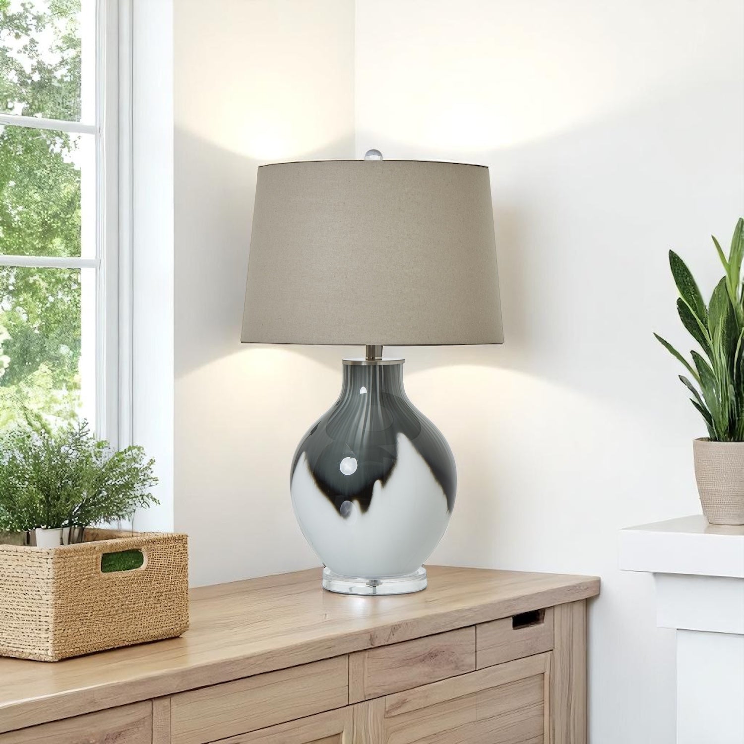 Grey and white glass table lamp with a beige shade on a wooden side table next to a window with plants.