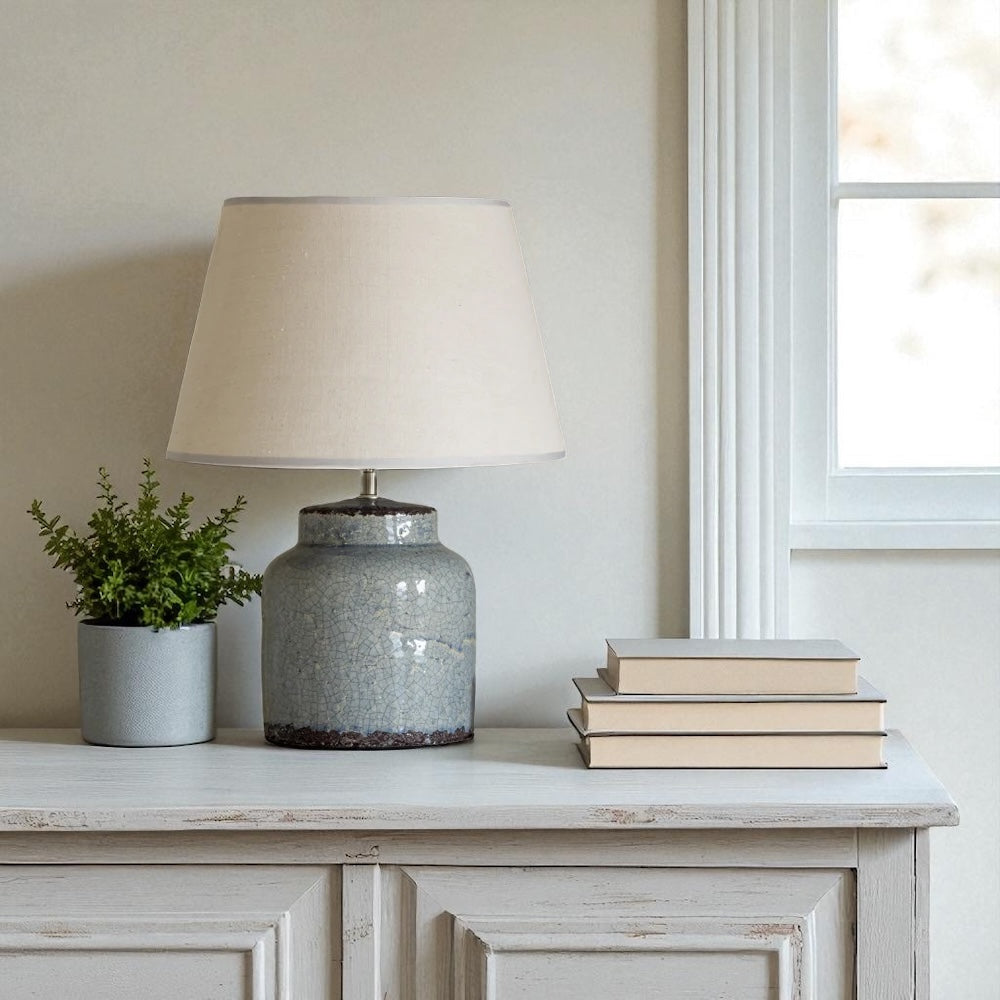 Blue ceramic lamp with cream lampshade on a wooden surface next to books and a small plant.