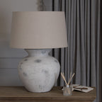 White rustic table lamp with beige shade on a wooden surface next to grey curtains