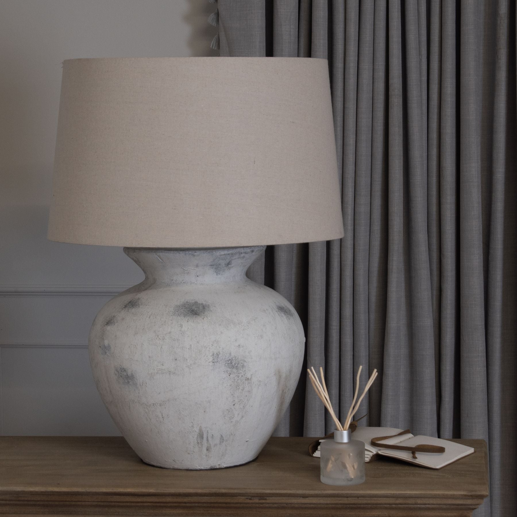 White rustic table lamp with beige shade on a wooden surface next to grey curtains