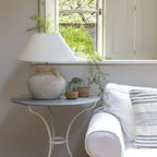 Decorative lamp on a small table next to a white sofa with a striped pillow, in a room with a window and plants.