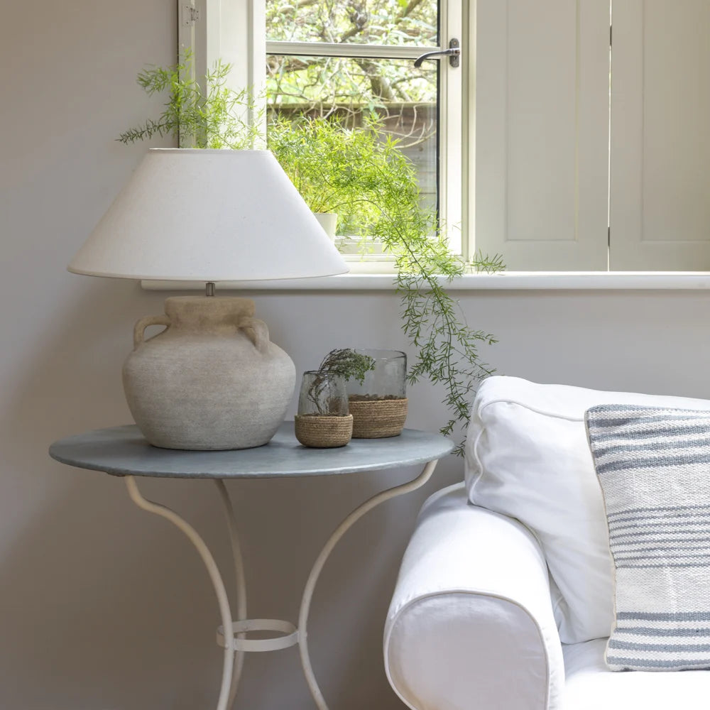 Decorative lamp on a small table next to a white sofa with a striped pillow, in a room with a window and plants.
