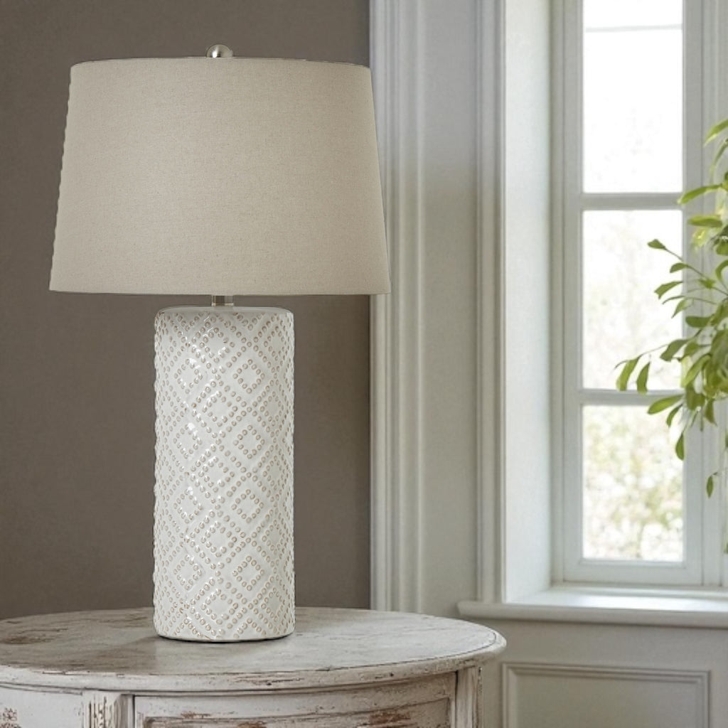 Table lamp with textured white base and cream shade on a wooden table near a window.