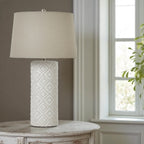 Table lamp with textured white base and cream shade on a wooden table near a window.