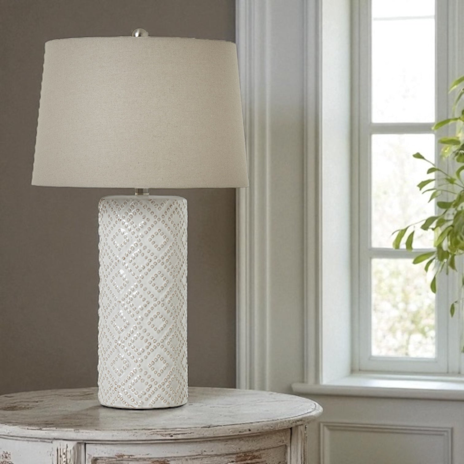 Table lamp with textured white base and cream shade on a wooden table near a window.