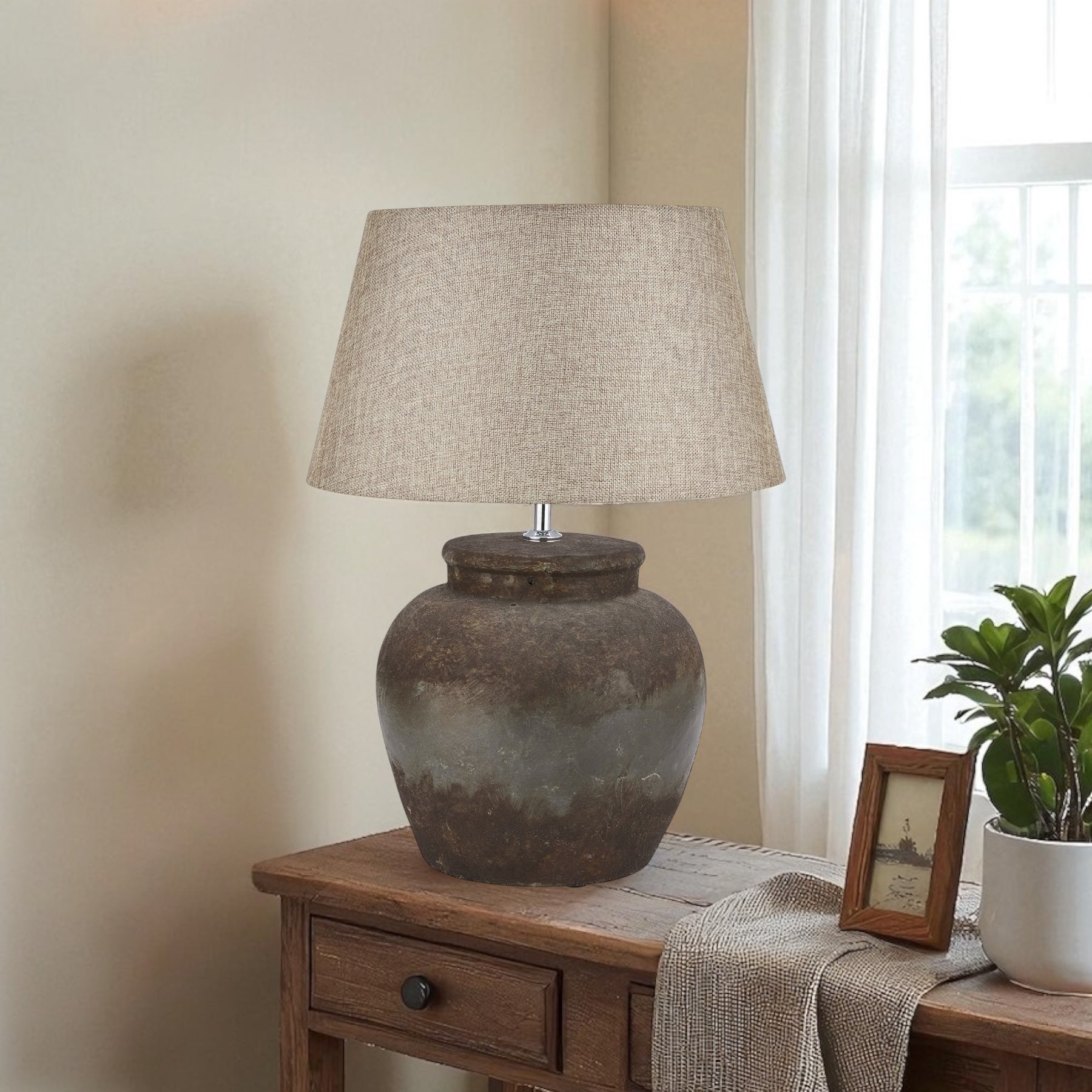 Hand painted stone effect table lamp with a linen shade on a wooden table next to a plant and photo frame.