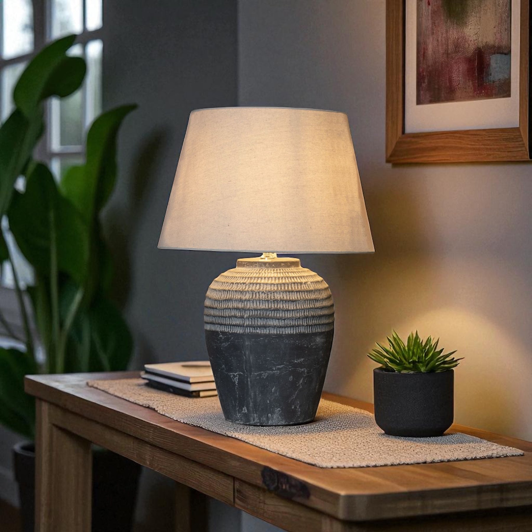 Table lamp with textured base and beige shade on a wooden table with a plant and books in the background.