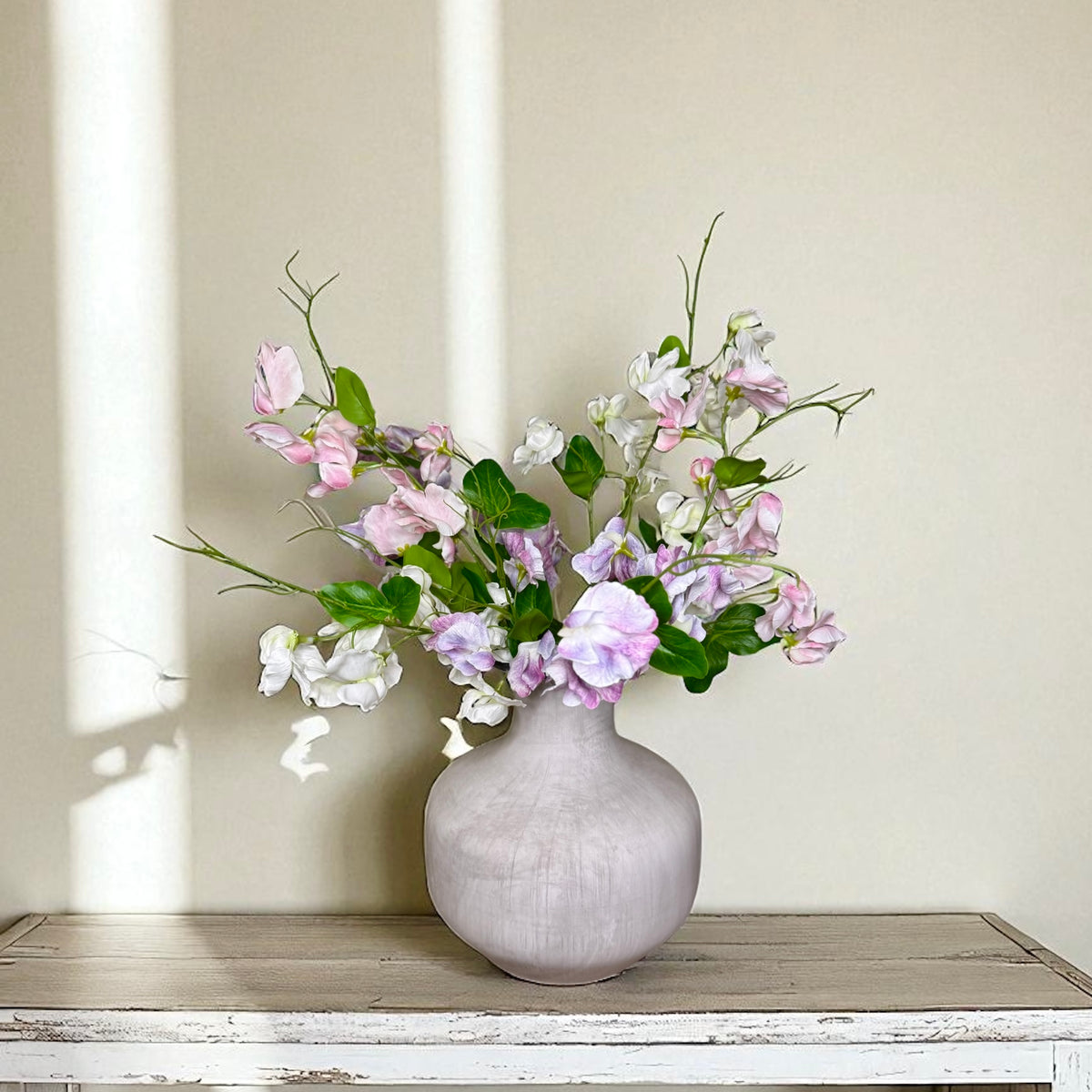 Gray textured vase with pink, purple and white flowers on a wooden surface against a beige wall.