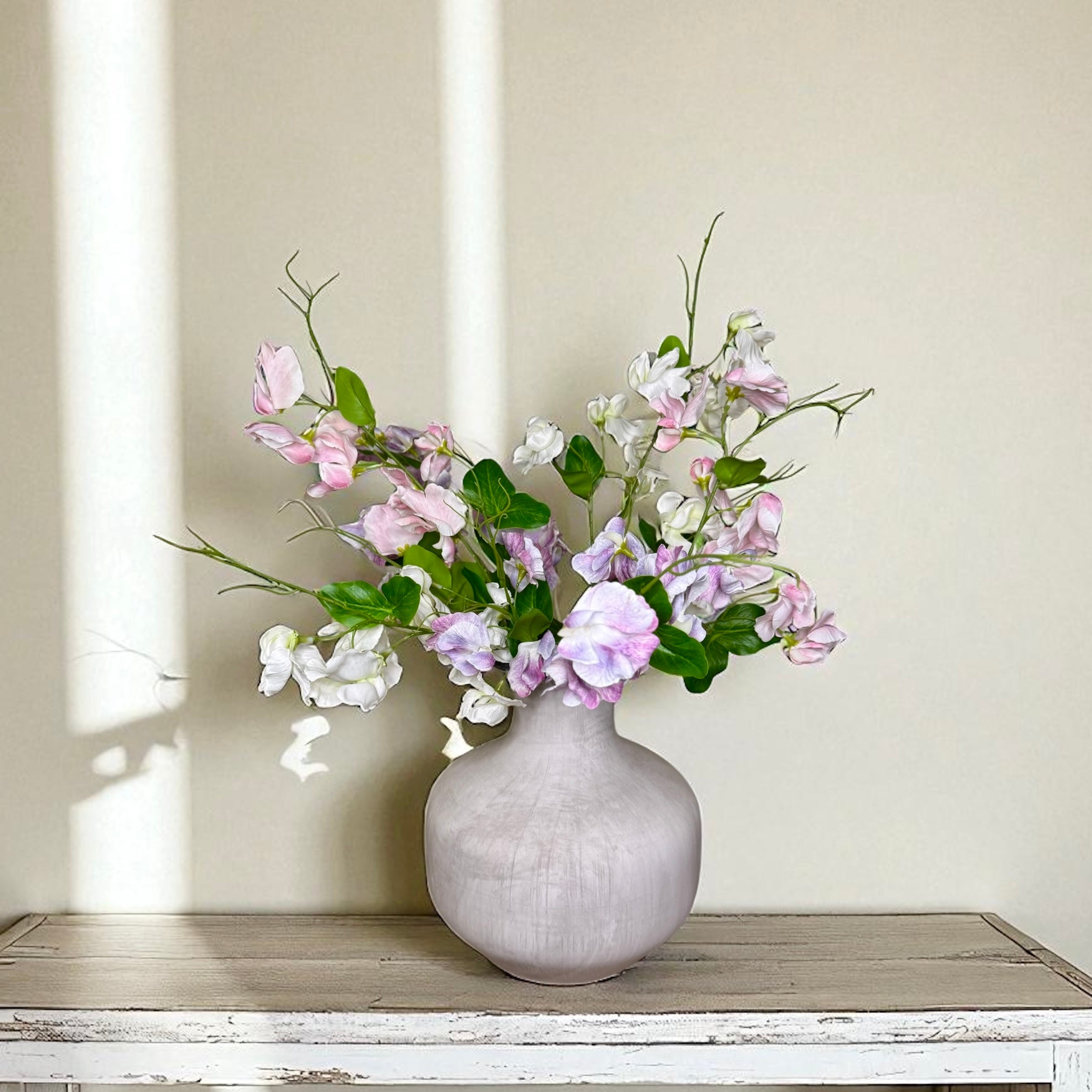 Gray textured vase with pink, purple and white flowers on a wooden surface against a beige wall.
