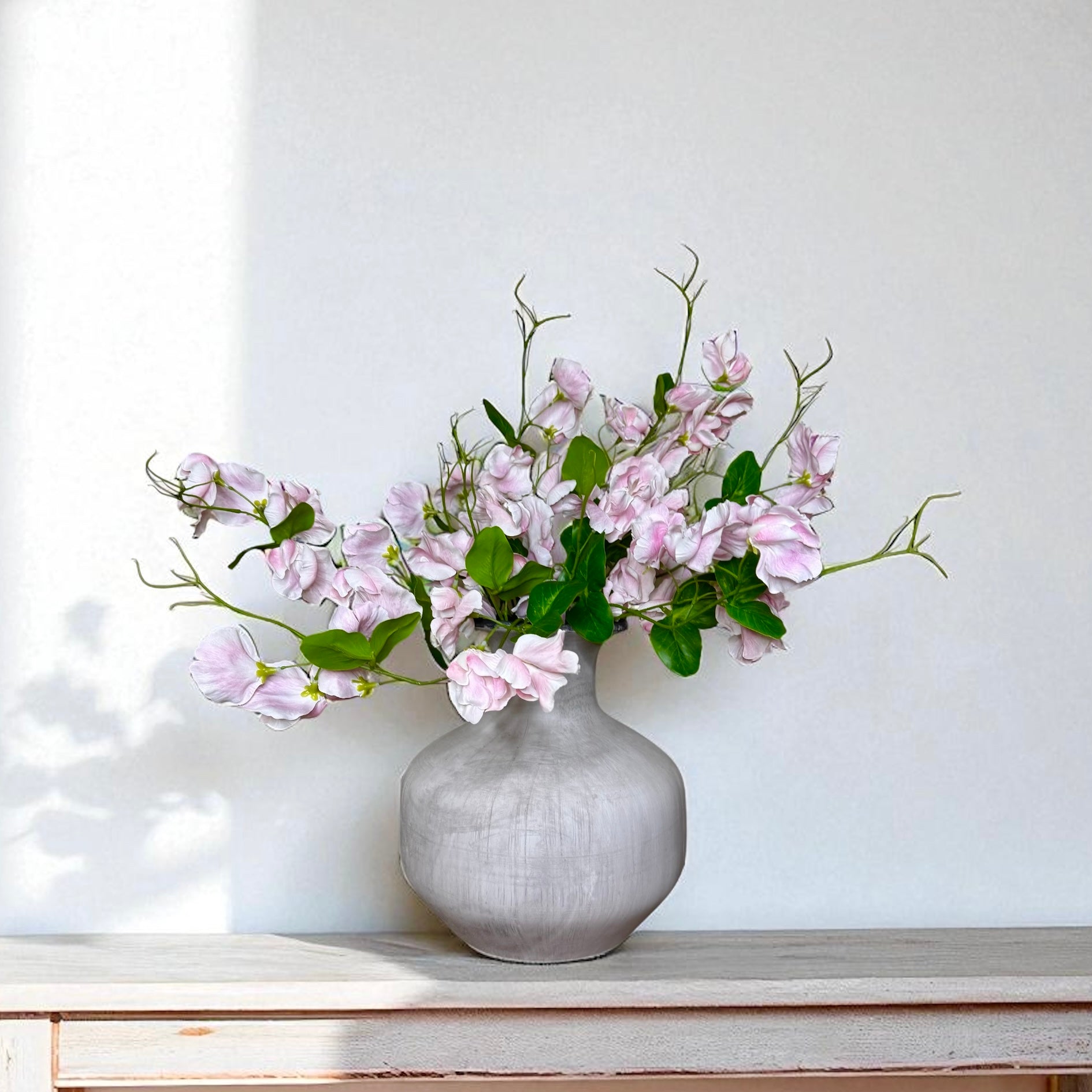 Grey vase with pink sweet pea flowers on a wooden surface against a white wall