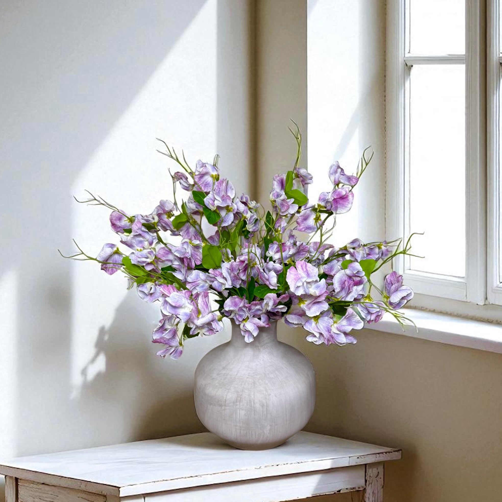 Light grey vase with purple sweet pea flowers on a wooden table by a window