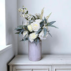 White floral arrangement in a grey vase on a wooden surface with a light grey wall background