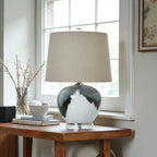 Decorative lamp with a beige shade on a wooden table near a window.