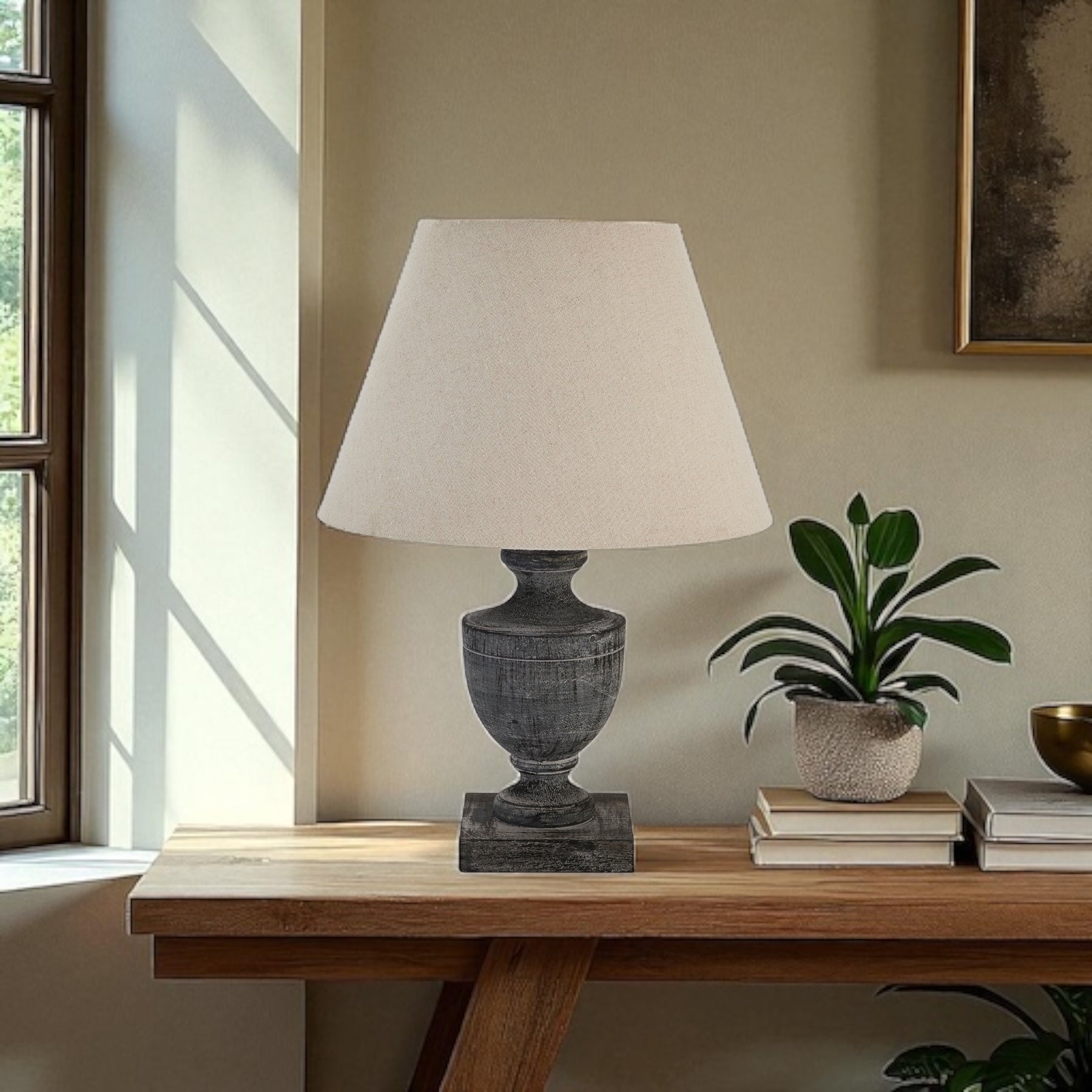 Table lamp with a white shade on a wooden table next to a plant and books, in a room with a window.
