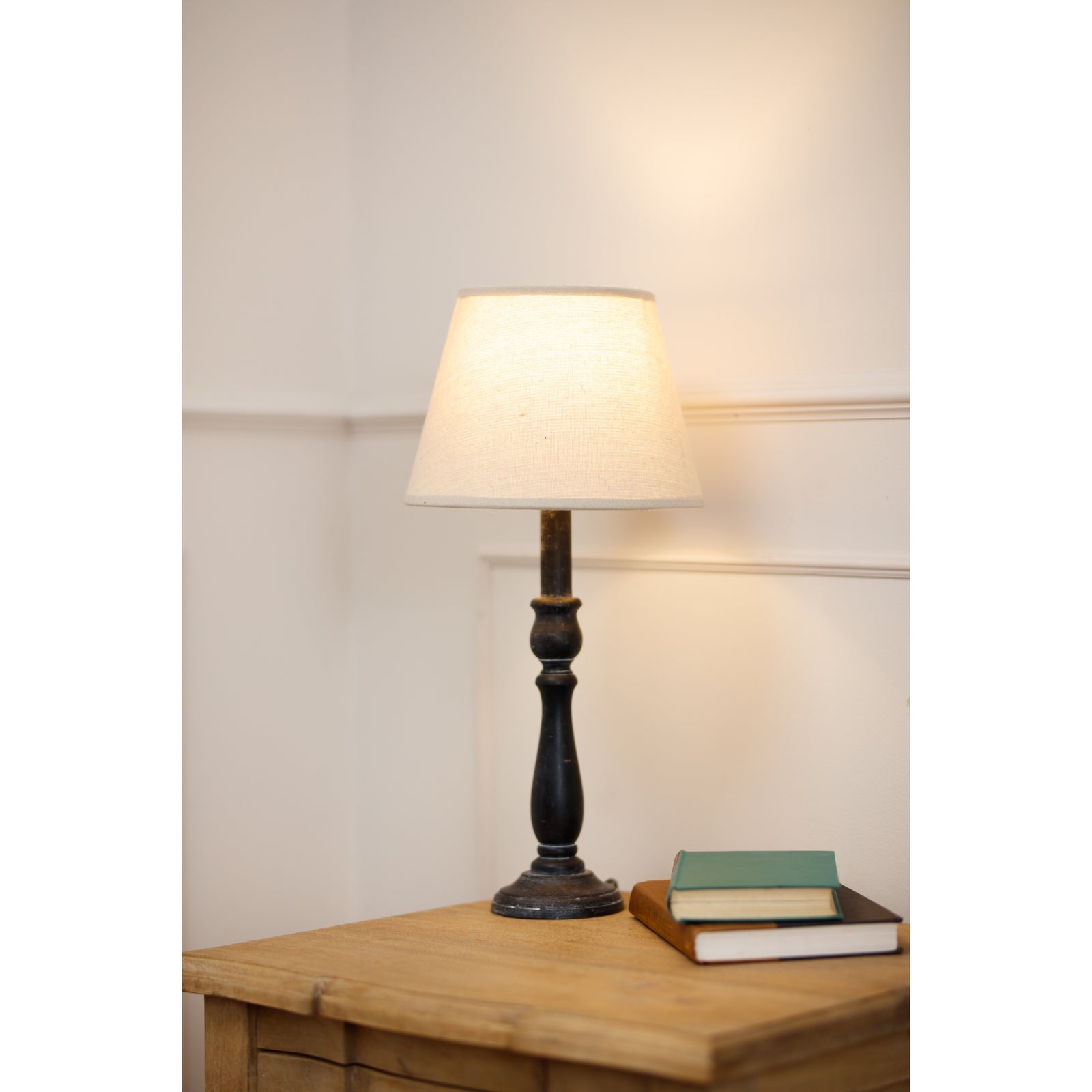 Verona Table lamp with a white shade on a wooden table next to a book against a neutral wall.