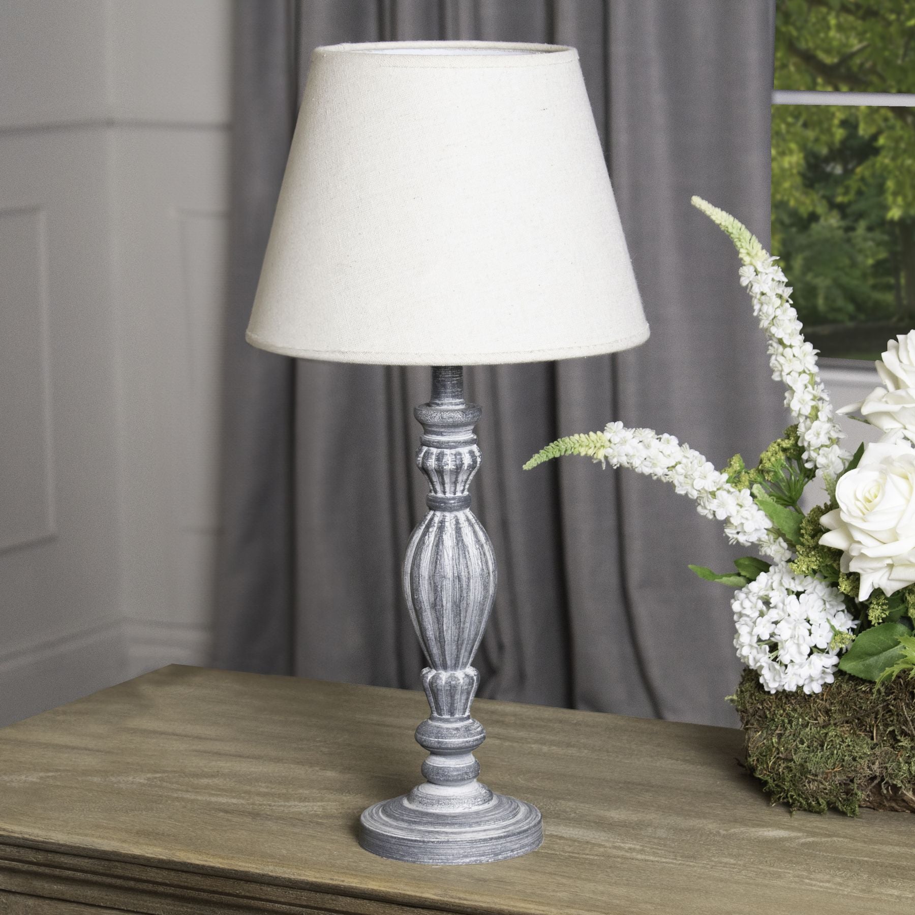 Table lamp with a cream shade on a wooden surface next to a floral arrangement.