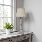 Table lamp with cream shade on a wooden side table next to a window with a plant.
