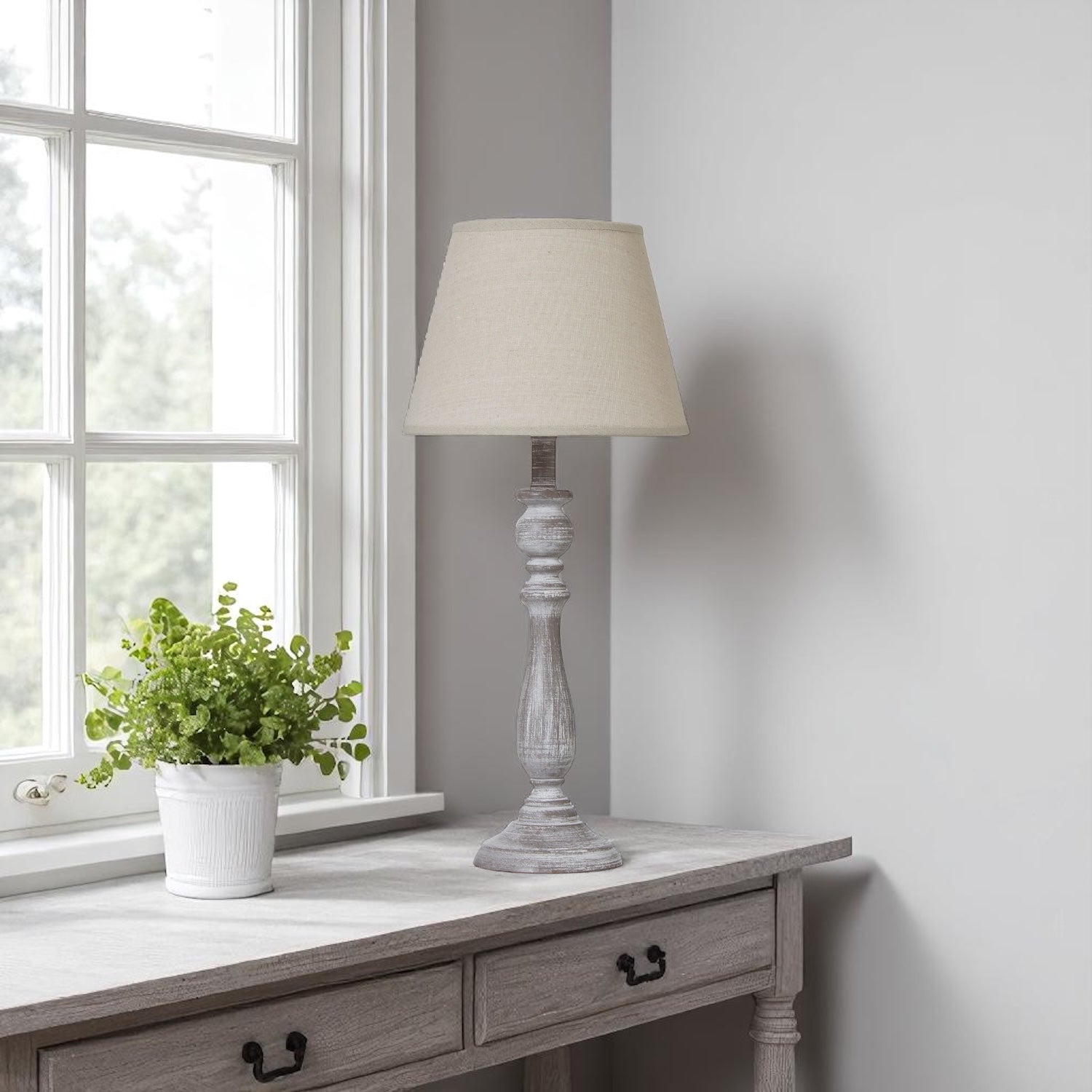 Table lamp with cream shade on a wooden side table next to a window with a plant.