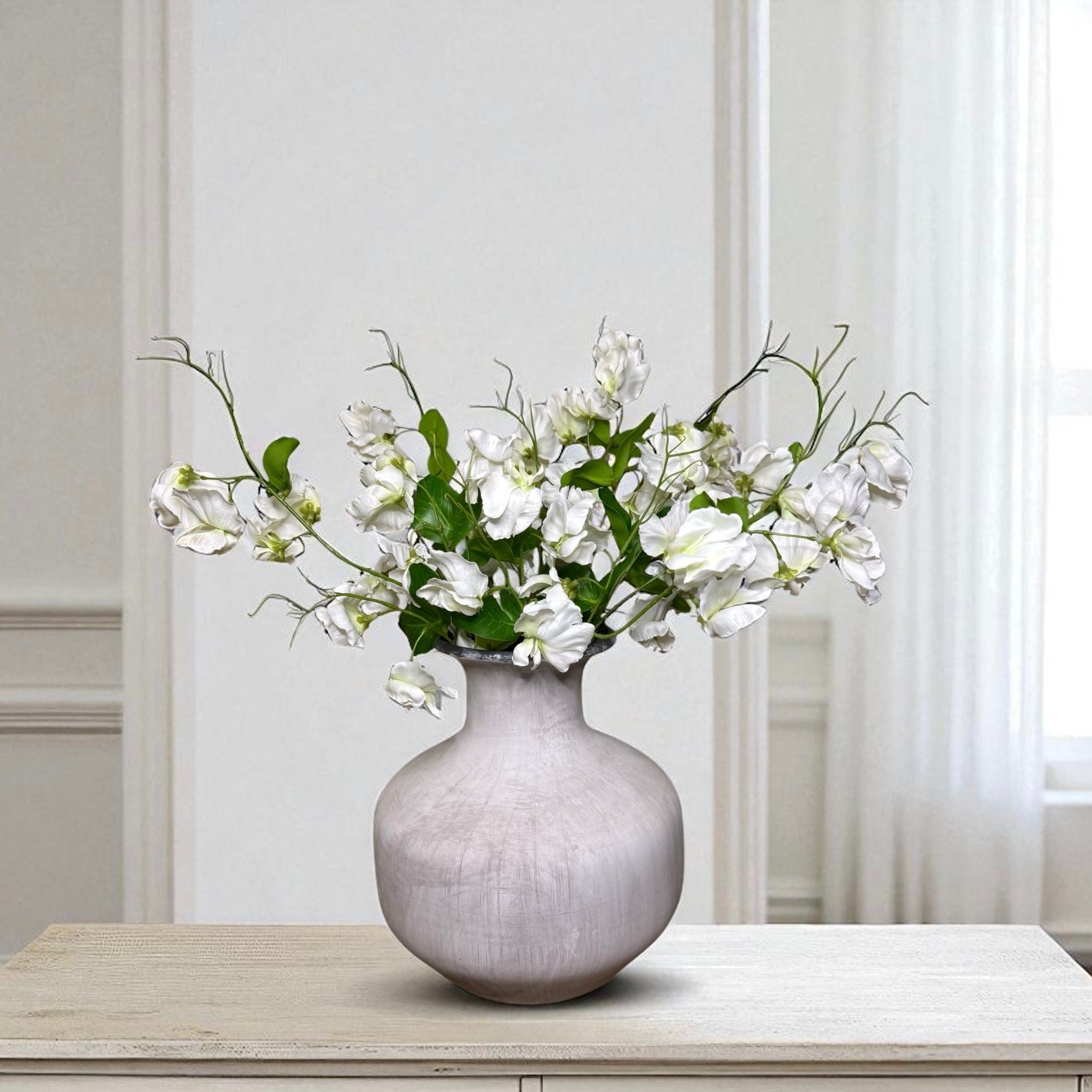 Grey vase with white sweet pea flowers on a wooden table against a light grey wall.