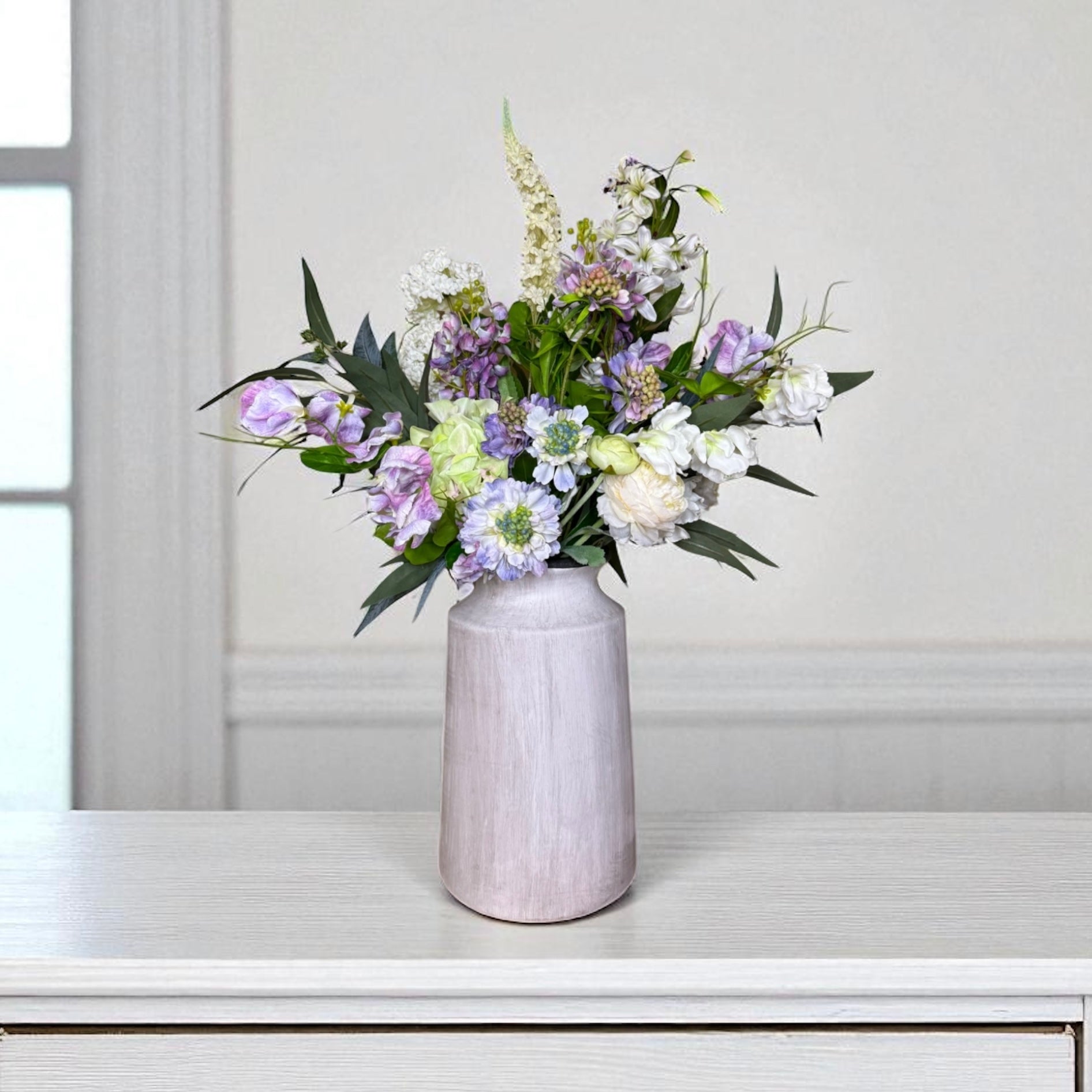 Purple and white floral arrangement in a grey vase on a white surface with a light grey wall background