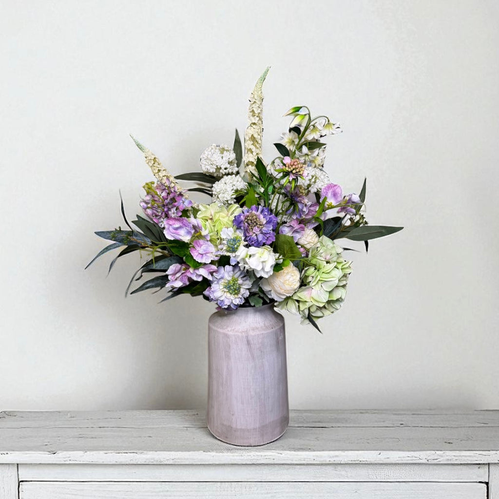 Purple and white floral arrangement in a grey vase on a white surface with a light grey background