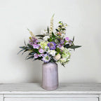 Purple and white floral arrangement in a grey vase on a white surface with a light grey background