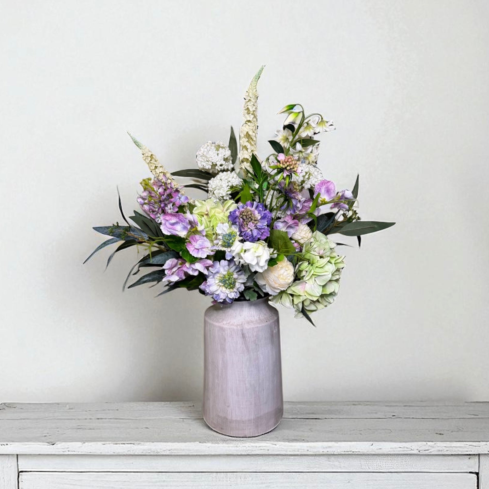 Purple and white floral arrangement in a grey vase on a white surface with a light grey background
