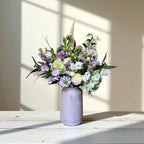 Bouquet of purple and white flowers in a grey vase on a wooden surface with sunlight casting shadows.