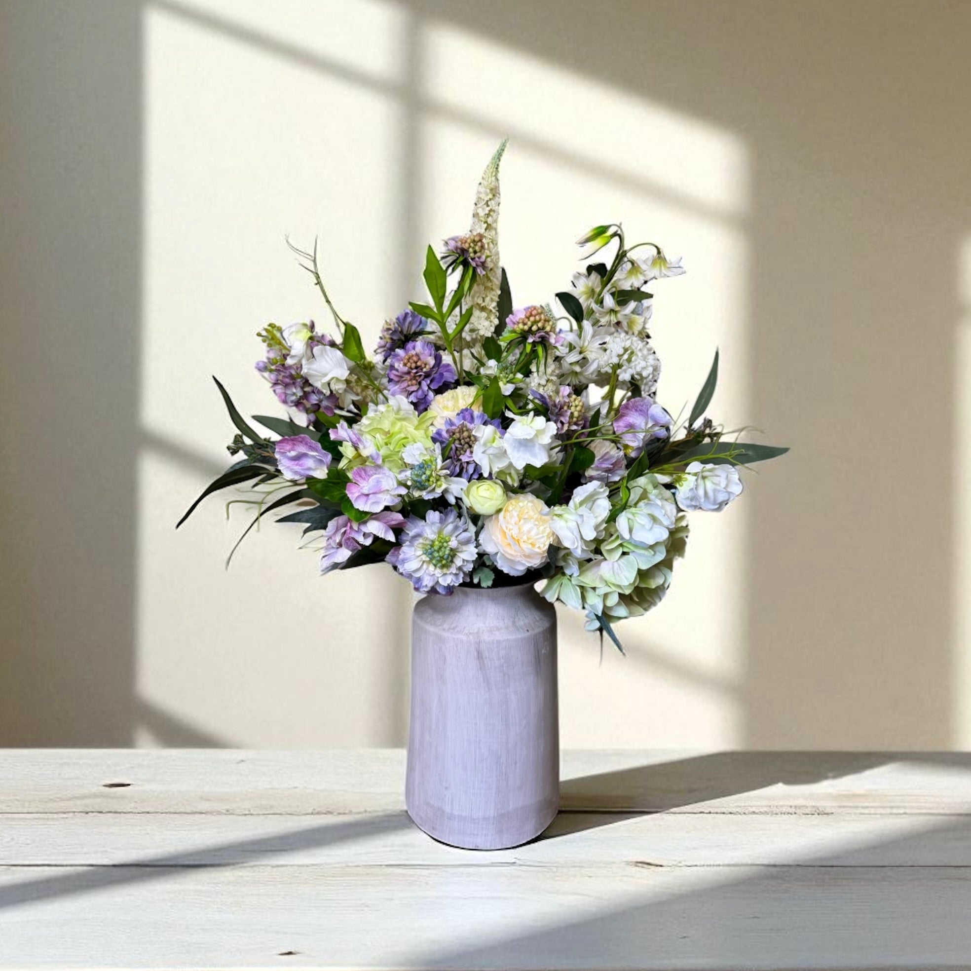 Bouquet of purple and white flowers in a grey vase on a wooden surface with sunlight casting shadows.
